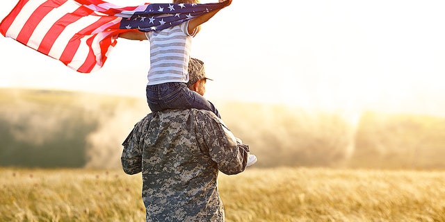A military servicemember holds a child on his shoulders as an American flag flies in the breeze. Said one Florida mom about her husband, "Regardless of what he finds, we built — and we will rebuild. We're Americans."